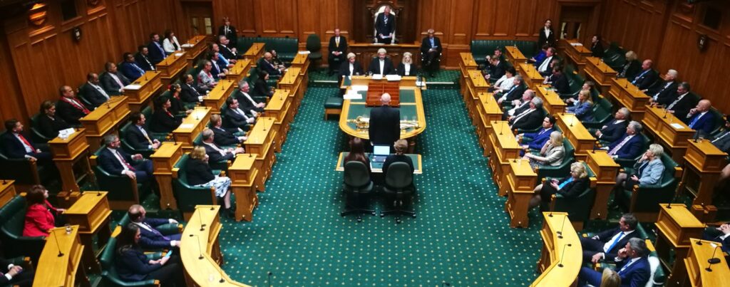Description: 
Photographie du Parlement de Nouvelle Zélande, construit en arc de cercle.
Les bureaux sont en bois clair, posés sur un tapis vert à pois blancs. Une cinquantaine de députés en costume noirs sont présents. 

Au centre de la scène, le speaker et le jury auditionnent un individu. Ils portent des perruques blanches à bouclette. Deux sténographes prennent des notes.