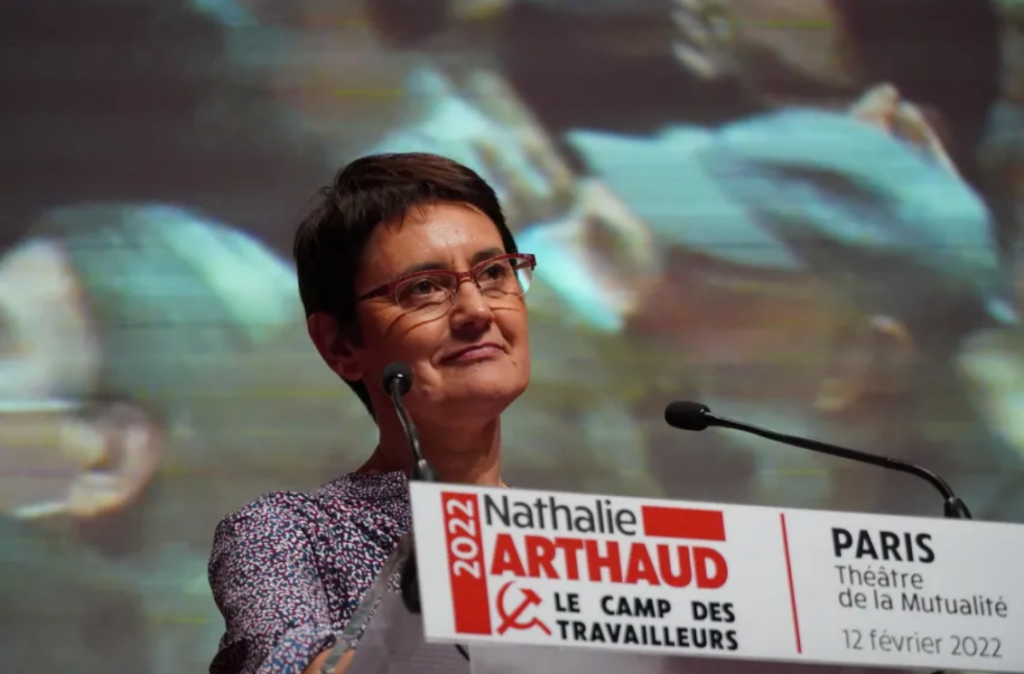 Nathalie Arthaud, candidate à l'élection présidentielle de 2022, à son meeting du 12 février 2022 à Paris. Elle pose derrière un pupitre et devant un écran.