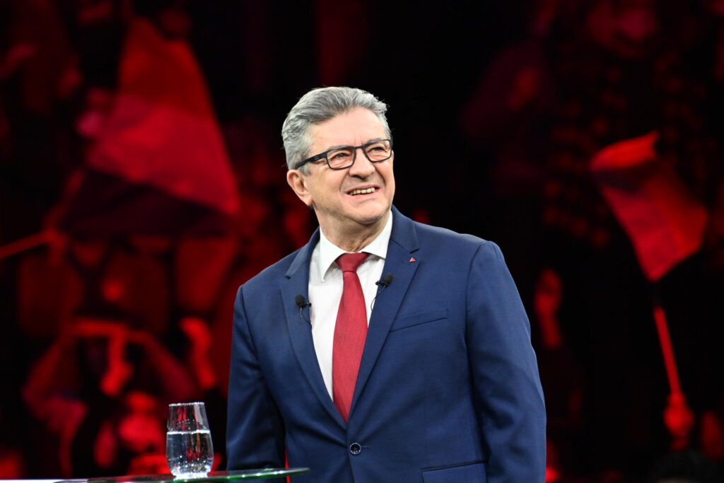 Au premier plan, Jean-Luc Mélenchon, candidat de La France Insoumise à l'élection présidentielle de 2022. Il est en meeting à Montpellier le 13 février 2022, souriant, en costume. À l'arrière plan, des militants flous agitent des drapeaux.